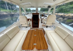 Interior of a luxury boat cockpit with cream leather seating, wooden table, and helm controls at the front, with water and rocks visible through the windows.