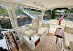 Interior of a luxurious boat cabin with cream captain chair at helm, beige seating, and a wooden table beside a teak deck, windows to water.