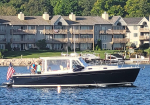 Motorboat sailing on blue water with several people aboard and an American flag at the stern, docked near a residential complex along the shoreline.
