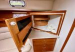 Wooden boat galley with an open cabinet; a microwave or oven inside and a white countertop with a cup rack, drawers below, and a porthole window above.
