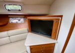 TV mounted in a teak cabinet inside a boat cabin, with beige leather seating and a small porthole window nearby.
