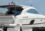 Side view of a white motor yacht docked in a marina, with a tinted cockpit window, sleek curved hull, and a fence with service signs in the foreground.