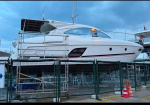 White luxury motor yacht on dry dock behind a green fence, with scaffolding and a cloudy blue sky.
