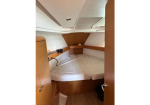 Wood-paneled boat cabin with a curved double bed, white sheets, and built-in storage along the walls; small hatch/vent above the bed and a doorway visible at the entrance.