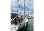 Sailboat moored at a marina with crew on deck and tall masts against a blue sky.