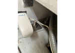 Close-up of a small white control box mounted on a metal surface, connected by white wires to a spring-loaded terminal, with a gray conduit nearby.