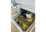 Open hatch on a sailboat deck showing coiled yellow rope, tangled gray lines, and a white storage box with a blue bin nearby.