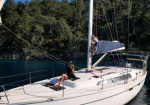 Two people relaxing on a white sailboat anchored on blue water with a tree-lined shore in the background.