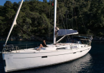 White sailing yacht anchored in calm blue water with two people sunbathing on the deck near a forested shoreline.