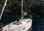 Two people aboard a white sailboat anchored in clear blue water near a forested coastline.