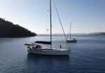 Two white sailboats anchored in a calm bay with a tree-covered coastline and distant hills; a person stands on the closer boat.