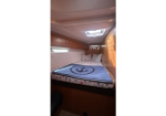 Compact boat cabin with a neatly made bed, blue anchor-pattern blanket, pillows, wooden cabinets overhead, and a skylight above.