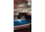 Boat cabin bedroom with a blue anchor-pattern bedspread, pillows, and curved white overhead storage; small window on the right.