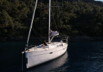 White sailboat on calm blue water near a tree-covered shoreline, with two people on the deck