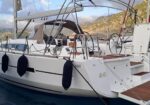 White sailboat moored in a marina, with black fenders along the hull, teak deck, and cockpit stairs, mountains visible in the background.