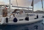 White sailing yacht moored at a dock with blue fenders along the hull and hills in the background.