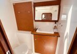Compact boat bathroom with wooden vanity, sink, and mirror; toilet in the corner and a soap bottle on the counter