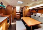 Inside a wooden yacht cabin: galley on left with stove and fridge, central dining table and cushioned bench seating, skylights above, warm wood paneling.