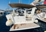 White sailboat at a marina viewed from the stern, with a wooden aft platform and two steering wheels under a canopy.