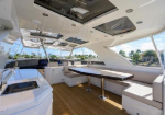 Luxurious yacht cockpit seating area with a long table, white cushions, teak deck, and overhead skylights; marina visible outside.