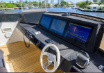 Yacht helm with three navigation screens, steering wheel, and throttle on teak deck; water canal and high-rise buildings visible in the background.