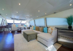 Luxurious yacht lounge with a beige sectional sofa, colorful cushions, and a green ottoman on a white rug, overlooking large windows.