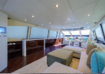Luxurious yacht interior: open salon with white sectional sofa, teal pillows, beige ottoman, wood floors, and large windows along both sides.