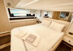 Modern yacht cabin with a large bed, white linens, and built-in wood cabinets along the wall near a window.