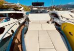 Front deck of a luxury motor yacht with cream sun loungers, docked between other boats on a sunny day at the marina.