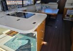 Galley on a boat with a white countertop, built-in stovetop, sink, and wooden drawers; a dining table and cushioned seating are in the background under windows with blinds.