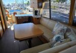 Sunlit yacht salon with beige sofa, nautical cushions, and a wooden dining table; marina and boats are visible through the windows.