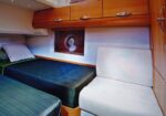 Boat cabin with two narrow twin beds, green-blue mattresses, wooden cabinetry, and a white cushioned seat nearby.