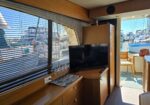 Yacht cabin interior with a flat-screen TV on a wood cabinet and striped window blinds overlooking moored boats. (Informative)
