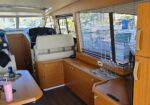 Interior of a luxury boat cabin with helm, cream seating, and wooden cabinets; blinds over large windows and wine glasses on a counter mat nearby.