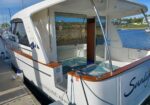 White motor yacht docked at a marina, rear deck with wood trim, open cabin door, and blue patterned deck rug visible.
