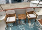Two wooden armchairs with white cushions and a small slatted table sit on a teal rug with a sea turtle motif on a boat deck.