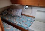 Twin berth cabin on a boat with seashell-pattern blue bedding, pillows, and wooden overhead storage; round porthole between white walls.