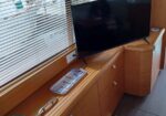 Television mounted on a wood cabinet inside a boat, facing a window with blinds and marina boats outside (TV off).