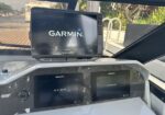 Boat cockpit showing three navigation screens: a Garmin chartplotter perched above two Raymarine AXIOM displays on a carbon-fiber panel.