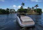 White luxury motorboat with beige cushions gliding on calm water near a tropical shoreline with palm trees and houses in the background.