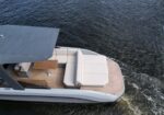 Top-down view of a white motorboat with beige cushioned seating and a wooden table on a teak deck, gliding through dark water.