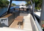 Open-air boat cockpit with beige cushioned seating, a teak table, and helm in view along a canal marina.