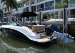 White and dark blue motorboat docked at a marina with a beige cockpit and black canopy, beside a modern building and palm trees.
