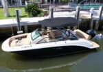 White and black motorboat with beige seating docked at a marina, with a dark canopy and teak deck area visible by a pier and glass-waced buildings in the background.