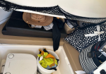 Boat cabin storage area with a straw hat, clothing, a polka-dot cushion, and two hats; a bucket of towels sits beside a portable toilet.