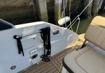 White boat deck with a stainless rail holding coiled black ropes, beige cushioned seating, and a small hatch, with a marina pilings backdrop.