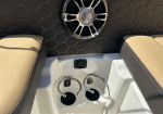 Boat cockpit area showing beige padded seats, a chrome speaker on a black hex-pattern wall, and two circular metal cup holders in a white console between the seats.