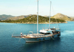 Twin-masted sailboat anchored in calm blue water near a rocky, green island and distant hills.