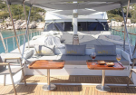 Luxurious yacht deck with white cushions, two small wooden tables, wine glasses and snacks, ocean view behind.