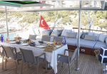 Outdoor yacht deck dining table set for a meal with plates, drinks, and lounge seating under a canopy; Turkish flag in the background.